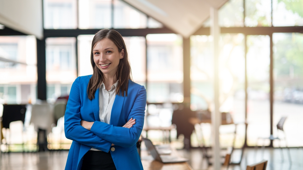 Remote Sales Rep at Join-Remote-Sales: A professional woman with a confident smile stands in a bright, airy office space, dressed in a smart blue blazer with arms crossed.